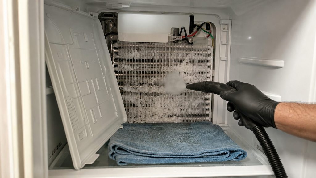 The freezer compartment with the rear panel off, evaporator fins packed with frost, defrost heater and sensor wiring above, a steam hose in hand and a towel catching the melt.