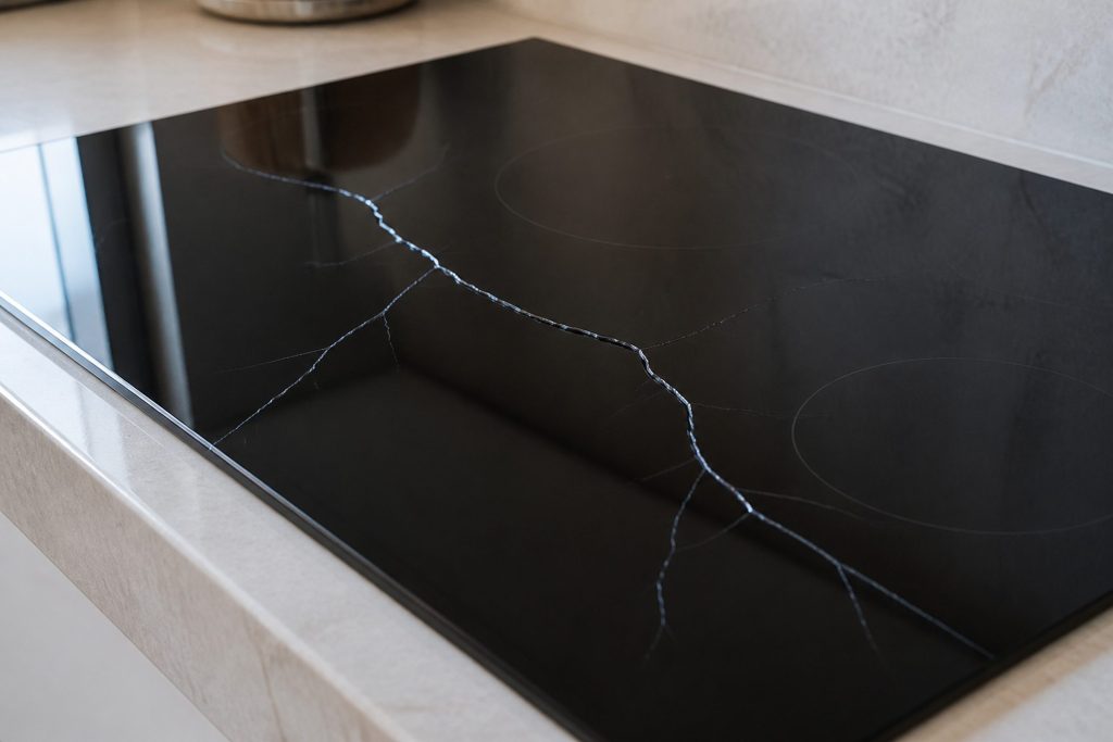 A dark ceramic glass cooktop surface with a long branching crack running across it, photographed at a low raking angle so the fracture catches the light, pale stone counter around it.