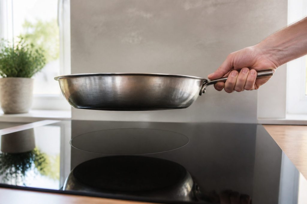 A heavy steel pan being lifted a hand's width off a black induction surface, the ring beneath it dark, a hand on the pan handle, bright kitchen daylight.