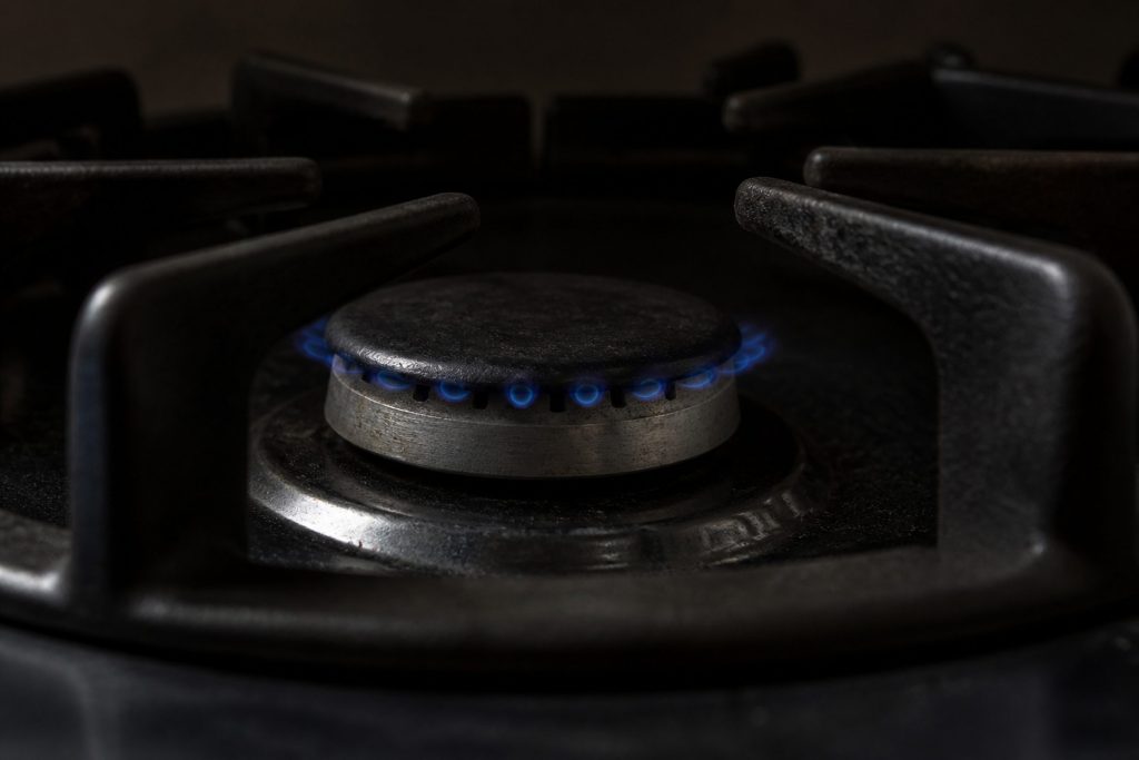 A gas burner turned right down, the crown of flame low, small and blue, several flame tips lifting away from the ports, photographed close in a darkened kitchen with a heavy pan support in the foreground.