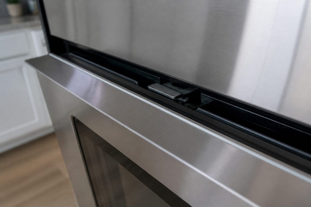 The top edge of a closed stainless oven door with the internal latch bar visible in the gap, photographed close and slightly from above in cool kitchen light.