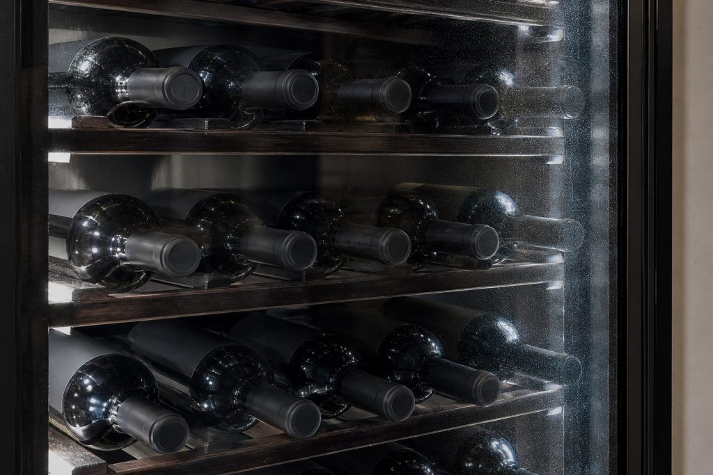 Bottles lying in dark timber racking inside a glass-fronted wine cabinet, the interior lit cool and even, condensation faint on the inside of the glass door.
